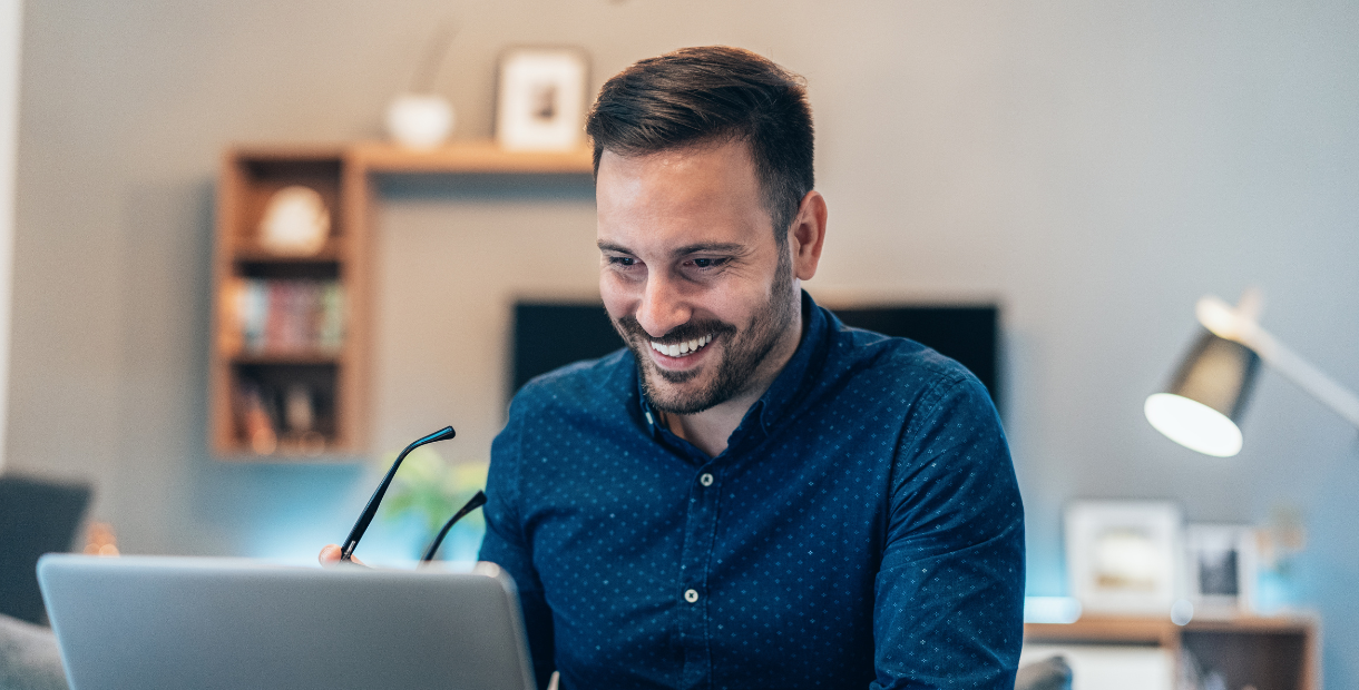 Person seated at a desk using a laptop, holding glasses, with shelves and a desk lamp visible in a home office background