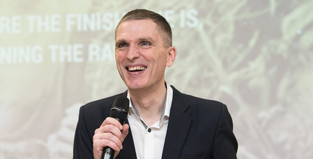 Mark Saxby in a dark suit holding a microphone while speaking to an audience at a presentation or workshop. A large screen with blurred text and graphics is visible in the background, creating a professional conference or training event setting.