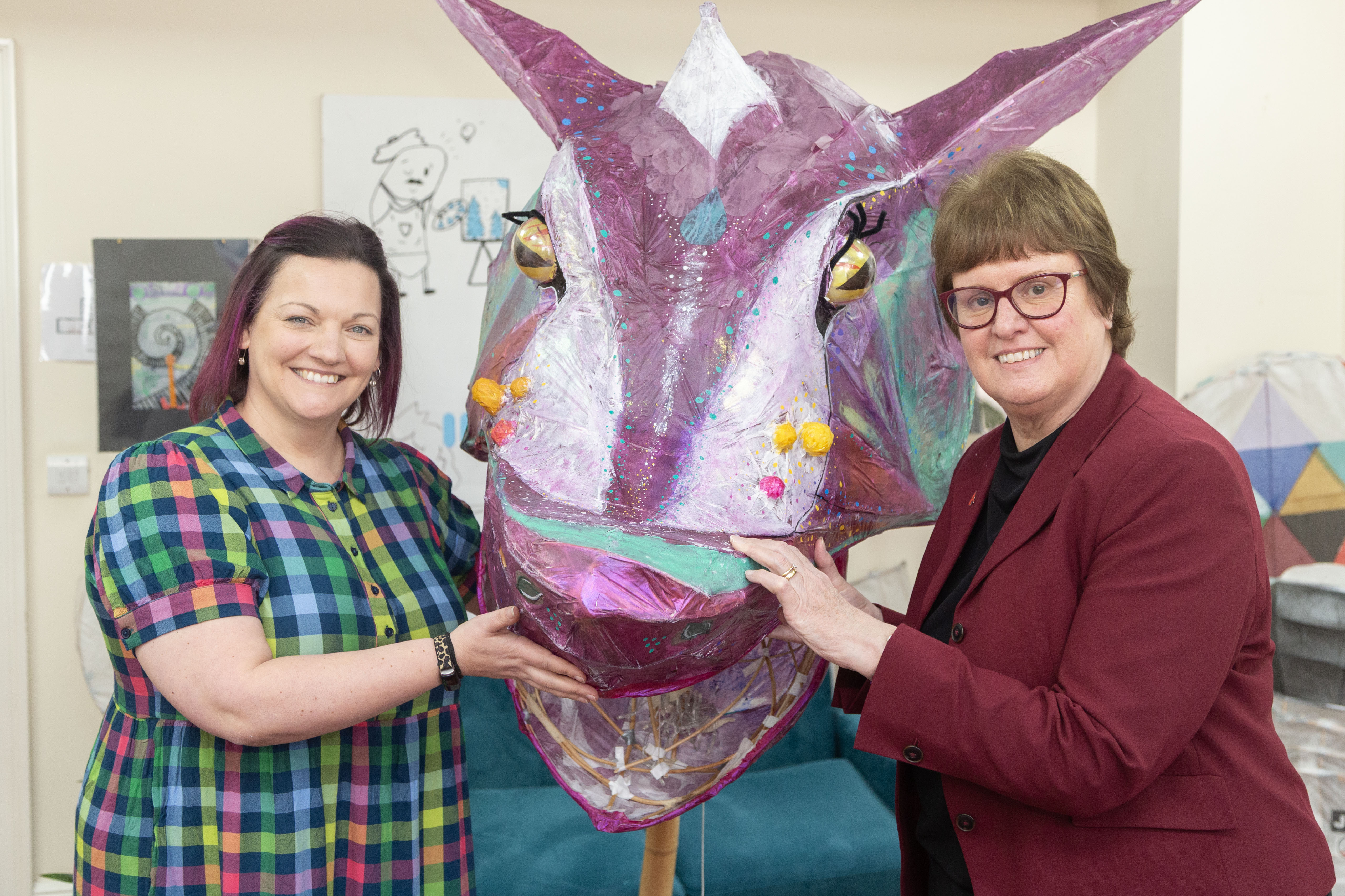 Emily Bowman from Junction Arts and Cllr Tricia Gilby stand indoors holding a large, colourful handmade dragon head sculpture between them. The dragon features bright purples, pinks and greens with painted details and pom poms. Artwork is displayed on the wall behind them.