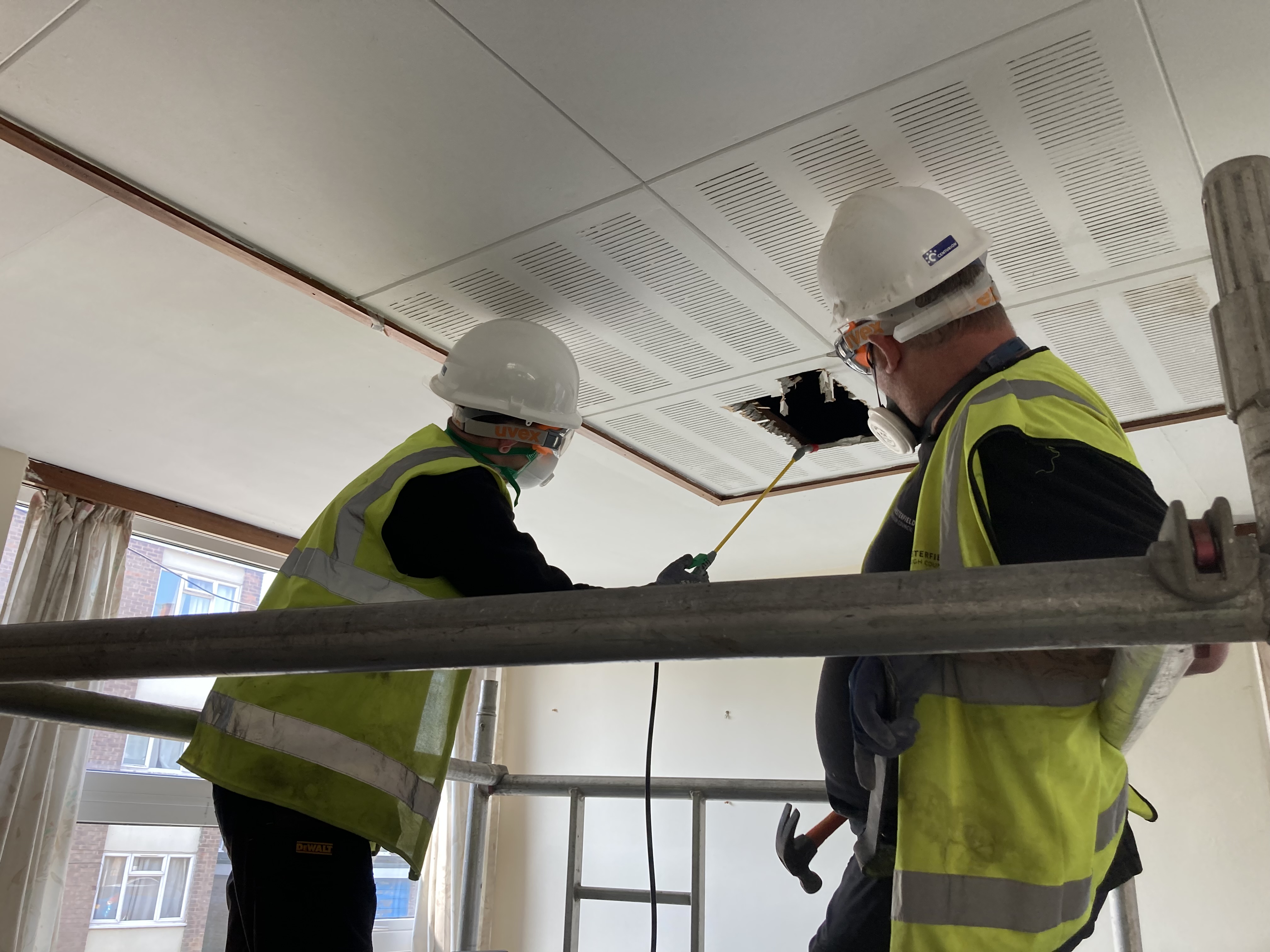 Two staff members stood on scaffolding wearing hard hats and hi-viz jackets. They are facing away from the camera and carrying out electrical work in the roof space.  